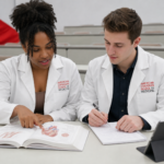 Two ACSOM medical students in white coats review anatomy notes and textbook diagrams together in a lecture hall, collaborating closely while studying cardiovascular anatomy with a notebook and tablet nearby.