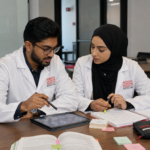 Two ACSOM students in white coats study together at a table, reviewing notes and medical scans on a tablet during a focused collaborative session in a modern campus workspace.