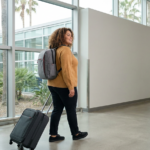 Incoming medical student walking through a bright, minimal airport terminal with a backpack and rolling carry-on, looking optimistic and prepared; the clean interior has light neutral walls and windows showing soft greenery outside. The subject wears comfortable travel clothes and is captured in a wide-angle full-body shot showing movement and purpose.