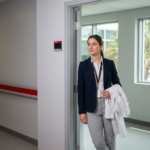 Medical student standing between a hospital ward corridor and an outpatient clinic hallway, pausing thoughtfully with a white coat draped over their arm and soft natural light from windows showing tropical greenery. The composition uses shallow depth of field to keep the student in sharp focus while the two clinical spaces are gently blurred.