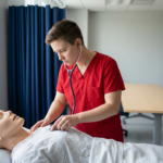 Medical student in red scrubs practices clinical assessment on a full-body mannequin in a clean simulation lab, surrounded by dark blue privacy curtains and soft lighting, conveying focused hands-on medical training.