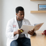 Medical student in a white ACSOM coat studies a licensure guide at a desk, holding a yellow highlighter beside a red mug in a bright office setting.