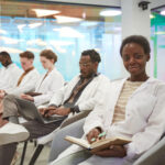 Portrait of young African American woman wearing lab coat and smiling at camera while sitting in row with multi-ethnic group of people in audience at medical seminar, copy space