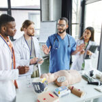 professional paramedic talking to diverse group of multiethnic students near CPR manikin, automated