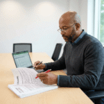 Prospective medical student at a clean conference-style desk reviewing tuition notes, scholarship paperwork, and a laptop spreadsheet while annotating with a pen under soft overhead lighting.