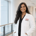 A smiling medical student in a white American Canadian School of Medicine coat stands in a bright hallway with windows, wearing black scrubs and hands tucked in pockets confidently indoors.