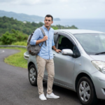A focused student stands beside a compact car on a scenic coastal road holding a backpack and looking prepared for the day, with soft, overcast light and a gently blurred background to keep the subject prominent.