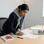 Student reviewing university transcripts and course notes at a laptop in a bright study office with a light gray wall and window greenery; focused and professionally serious academic moment.